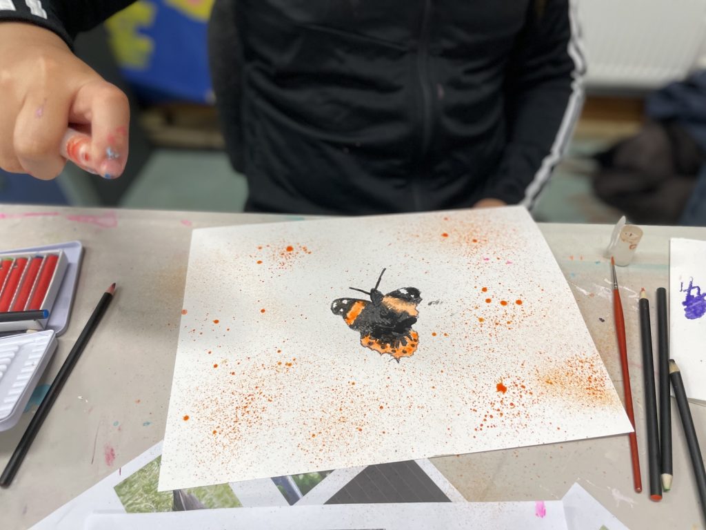 A photo of childs hands sparing orange ink onto a painted butterfly