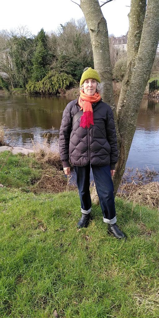 A photograph of a woman standing in front of a tree at the river's edge