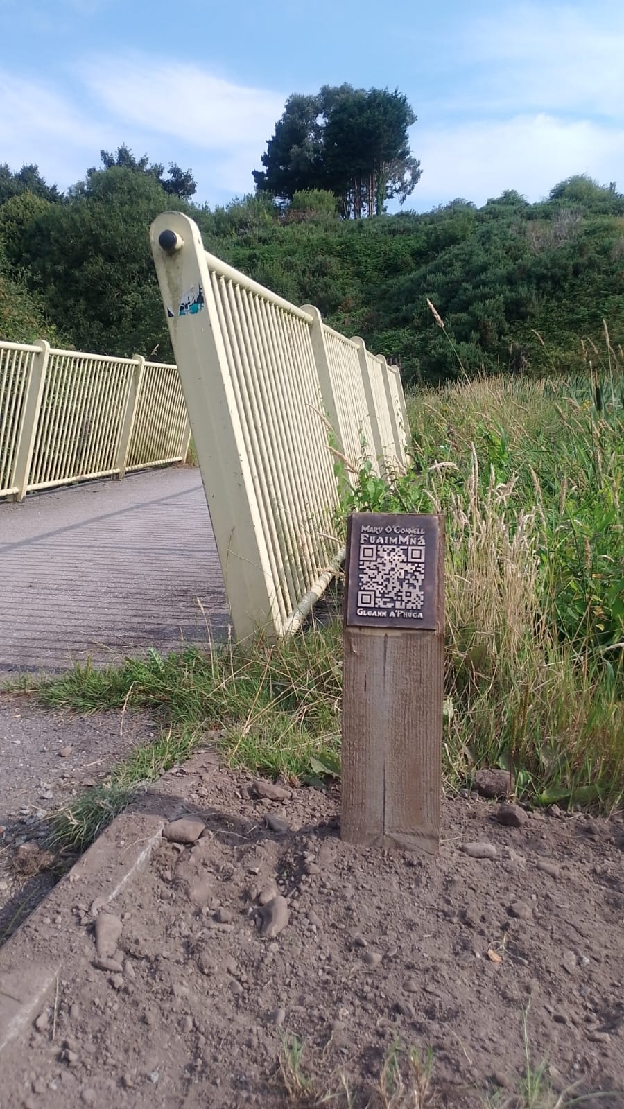 A photo of the railing of a bridge and a wooden post with a bronze QR code on it