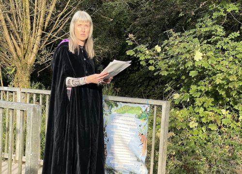 a photograph of a fair haireed woman holding a paper wearing a black claok and standing on a small stool near a bridge int a park