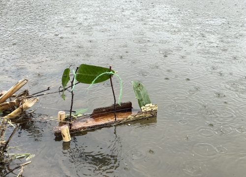 A photo of a toy boat made from bark and corks floating on water in the rain