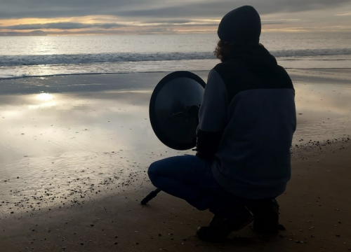 a photo of a man crouching on the shore at sunset with a recording device