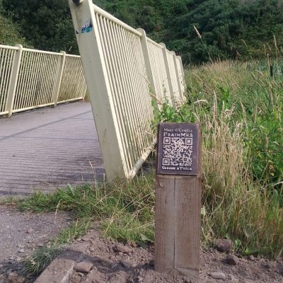 A photo of the railing of a bridge and a wooden post with a bronze QR code on it