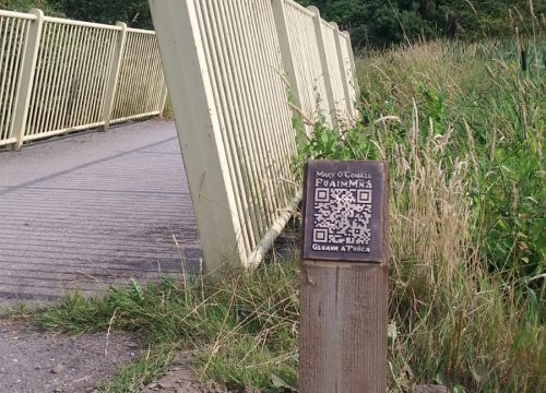 A photo of the railing of a bridge and a wooden post with a bronze QR code on it