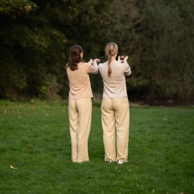 two women dressed in beige clothes with their backs to us holding cupped hands in a green park