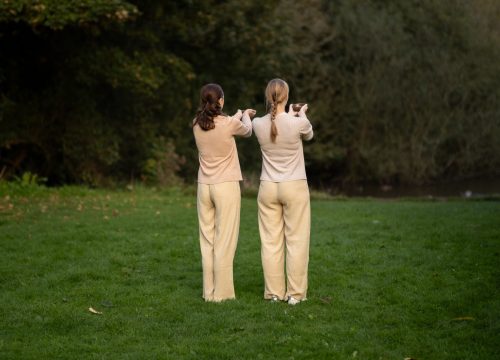 two women dressed in beige clothes with their backs to us holding cupped hands in a green park