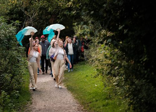 Two women dancers wearing beige and waving blue silk are followed by an audeience in a park